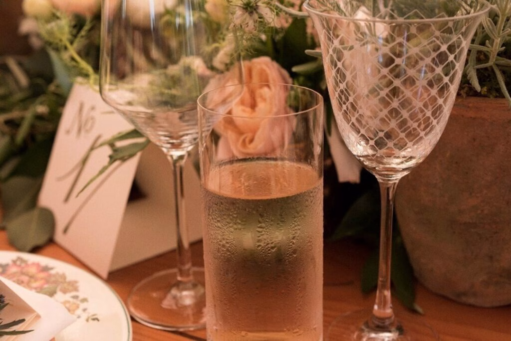 Close-up of an elegant retreat dinner table featuring a chilled glass of white wine with condensation, a lattice-patterned wine glass, a table number card, and warm pink roses.